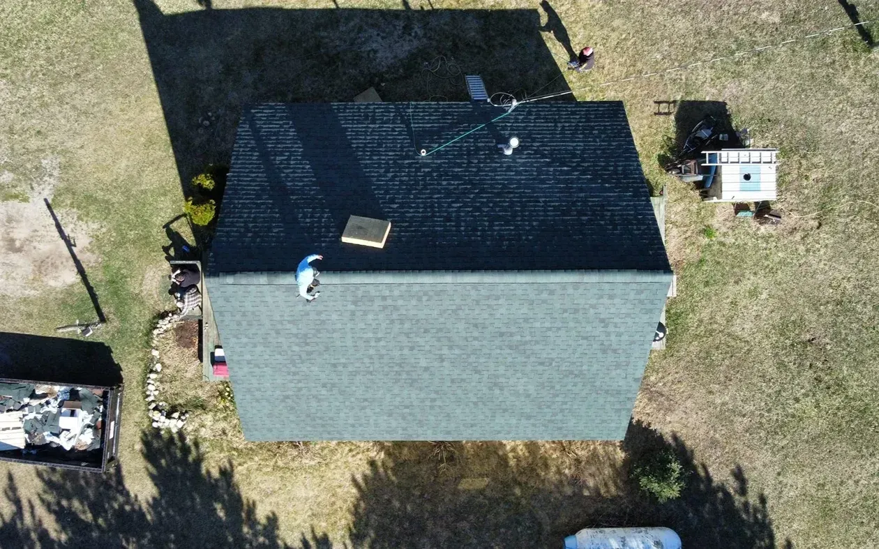 An aerial view of a house roof being repaired, with a person sitting on the shingles and construction debris below.