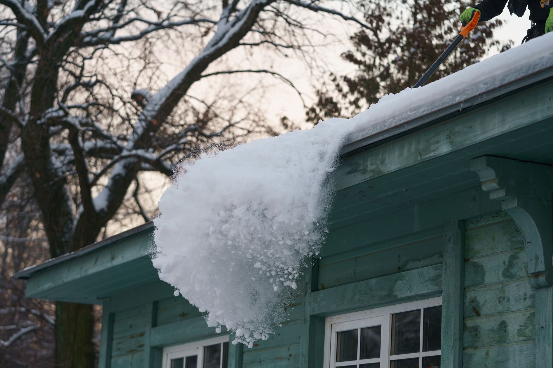A person clearing a large mass of snow off a teal-colored house roof with a long-handled tool during winter.