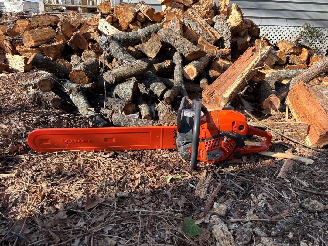 Orange chainsaw on the ground in front of stacked cut firewood outdoors.