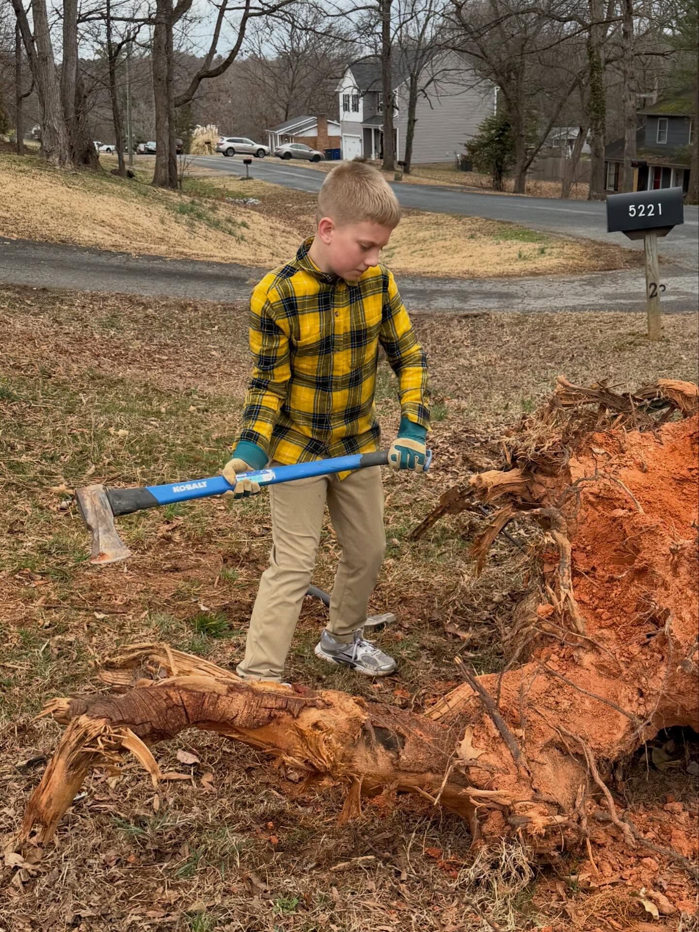 Person in a yellow plaid shirt uses a shovel beside a large orange dirt mound in a yard.