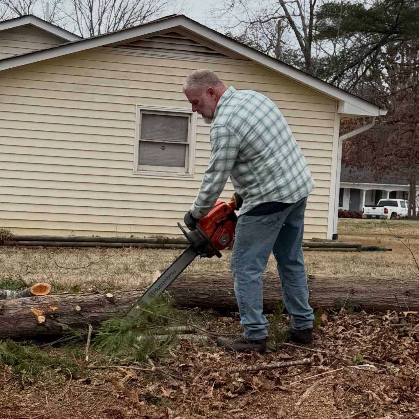 Man using a chainsaw to cut a fallen tree trunk in a yard beside a beige house