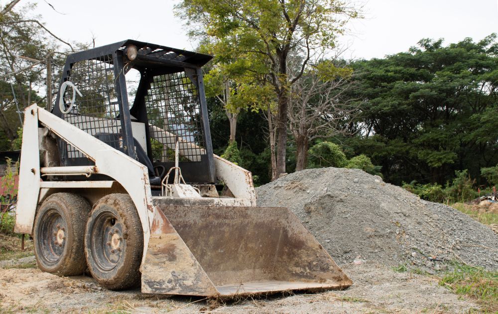 Skid steer loader parked beside a gravel pile on a dirt lot, with trees in the background