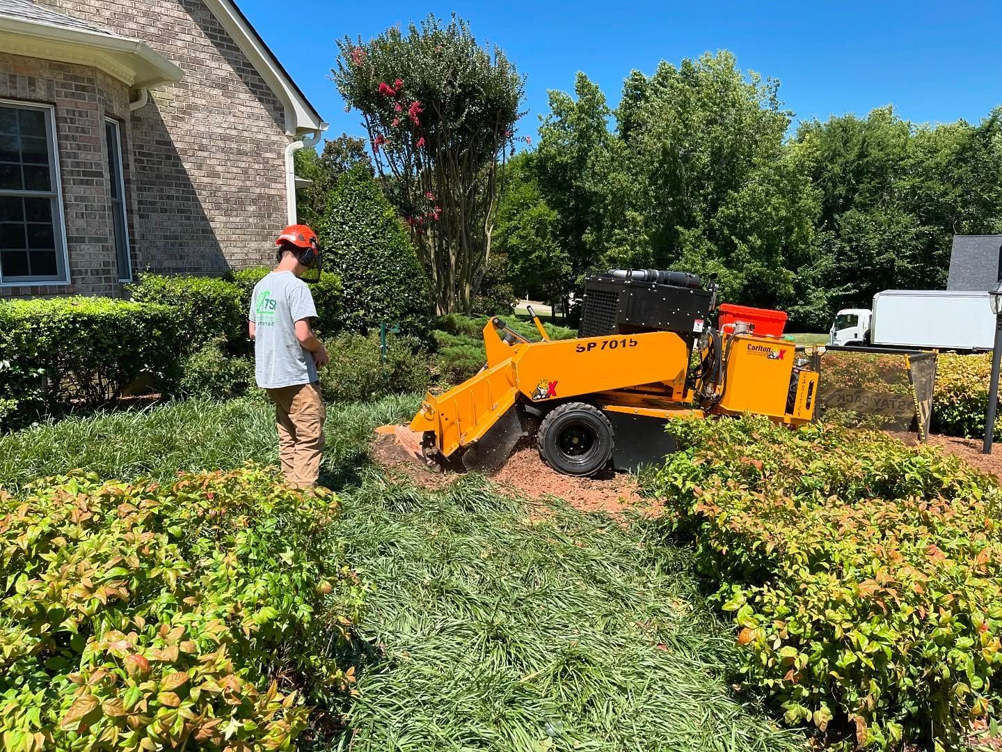 Worker standing beside an orange stump grinder in a landscaped yard near a stone house