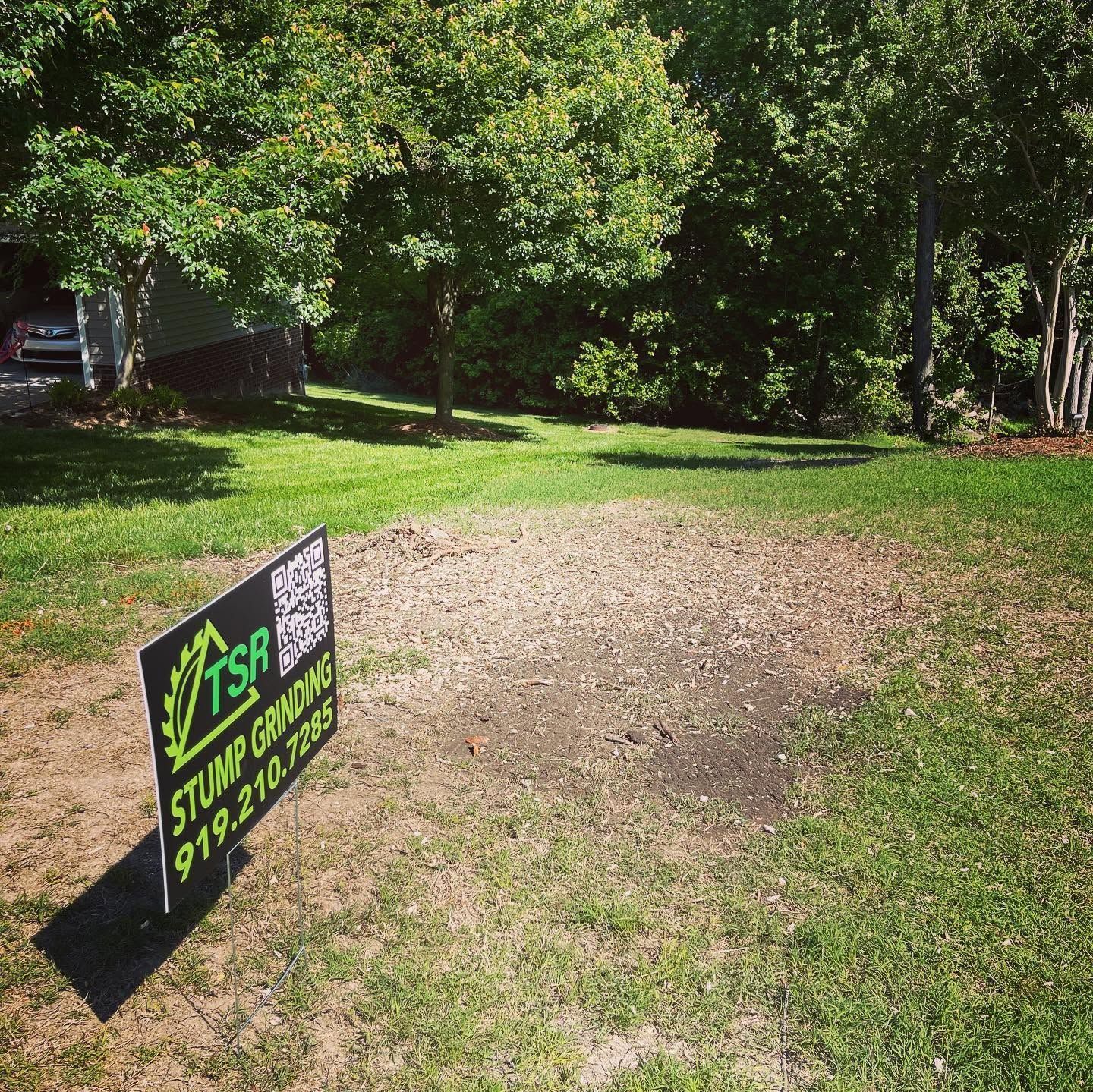 Yard sign for a stump grinding service on a sunny grassy lawn near trees.