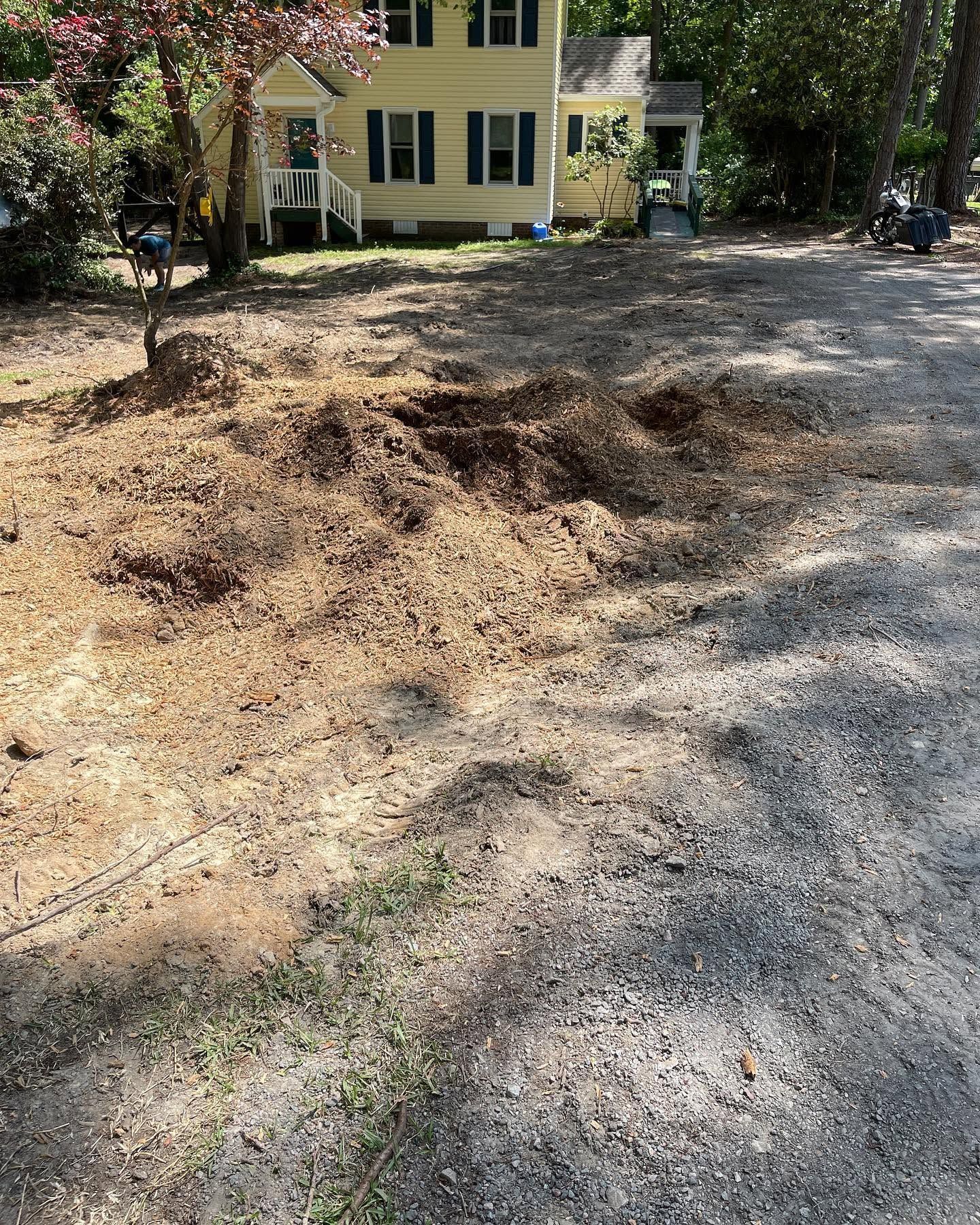 Gravel driveway with a large dirt mound in front of a yellow house and trees