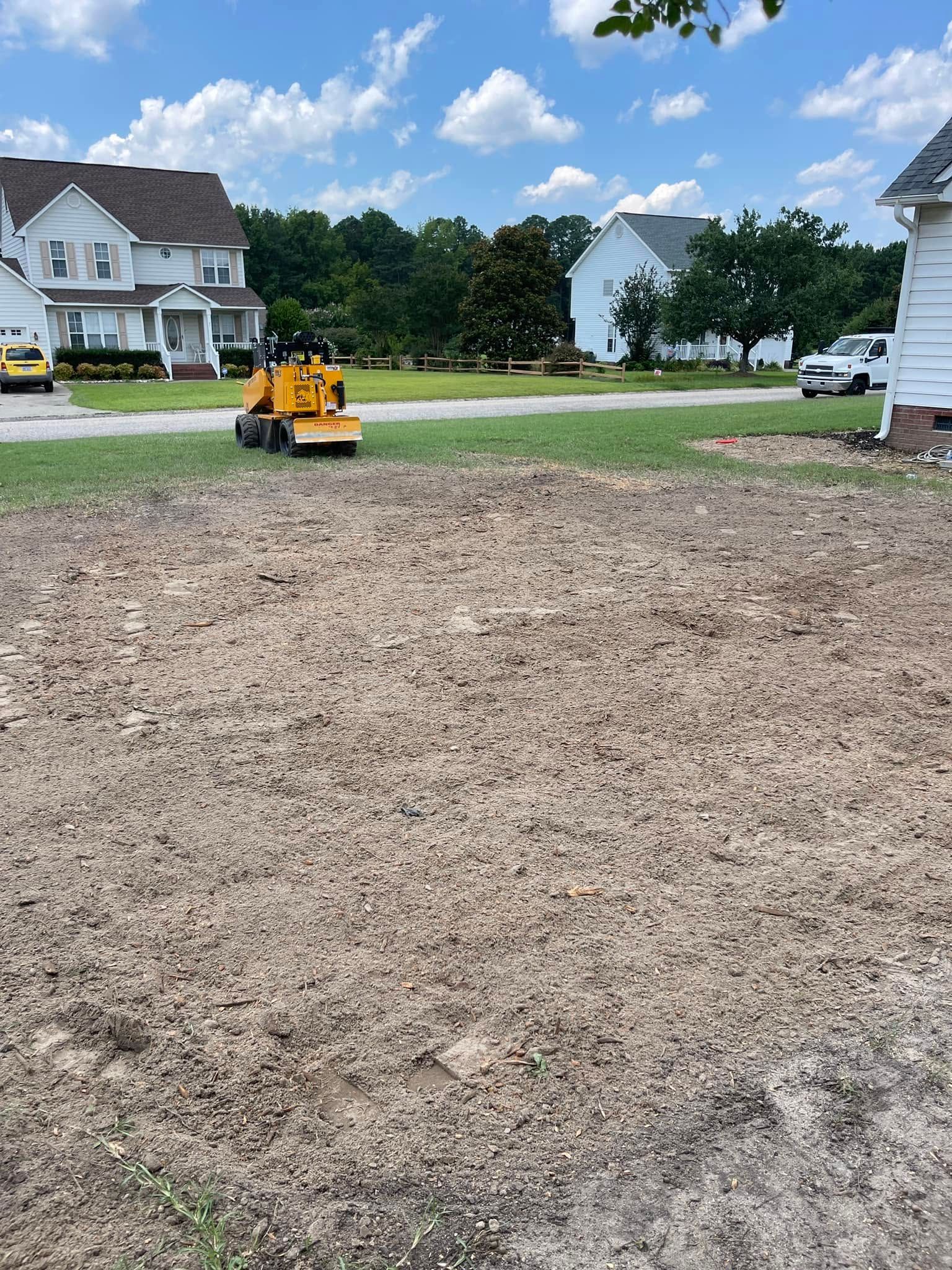 Yellow skid steer on a dirt lot in a suburban neighborhood with houses and trees in the background