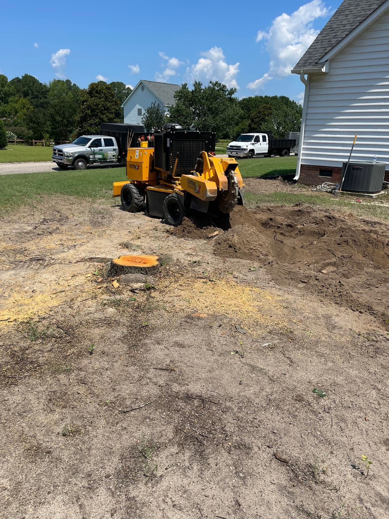 Yellow skid steer on dirt driveway near white house, with gravel and parked vehicles in the background.