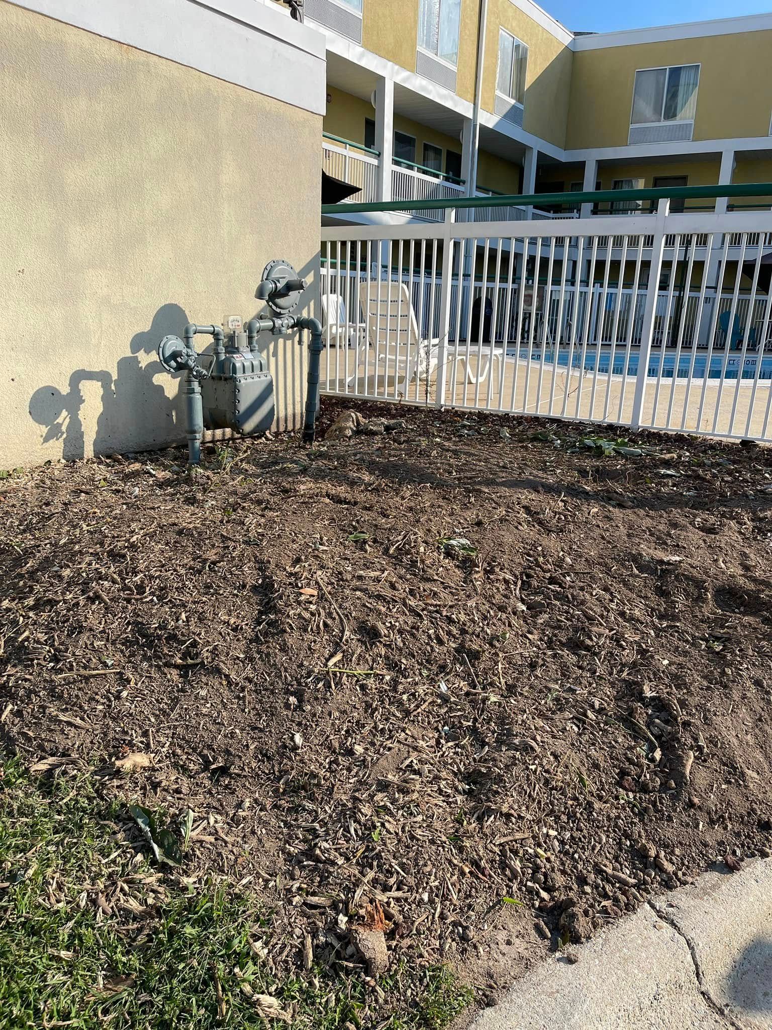 Mulched landscape bed beside a white fence and apartment building in bright sunlight