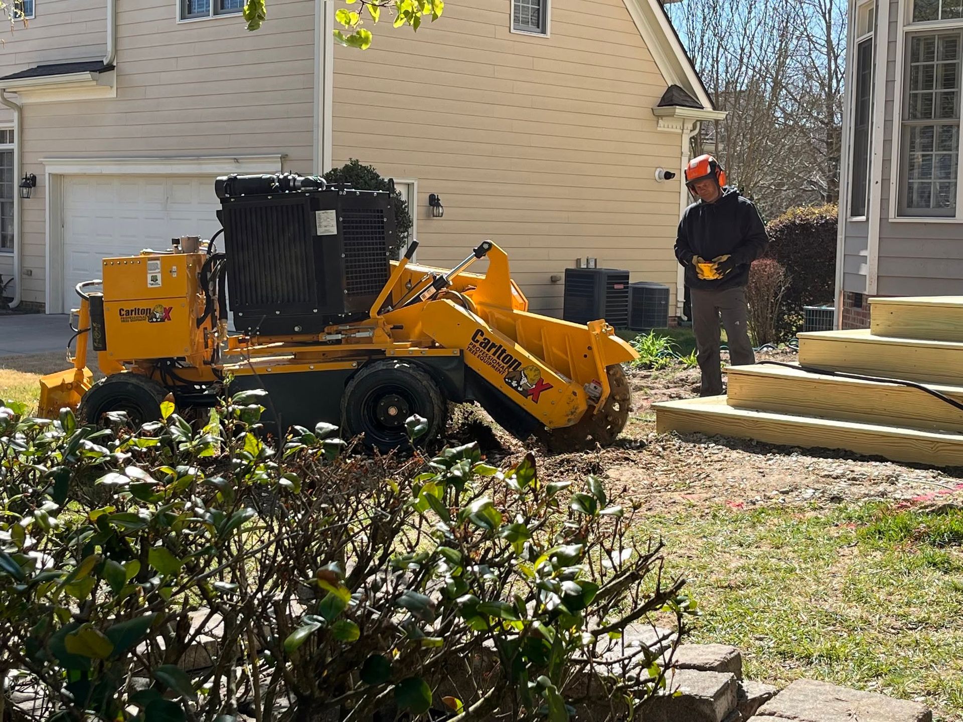 Yellow wood chipper beside a house, with a worker in a red hard hat standing near stacked lumber.