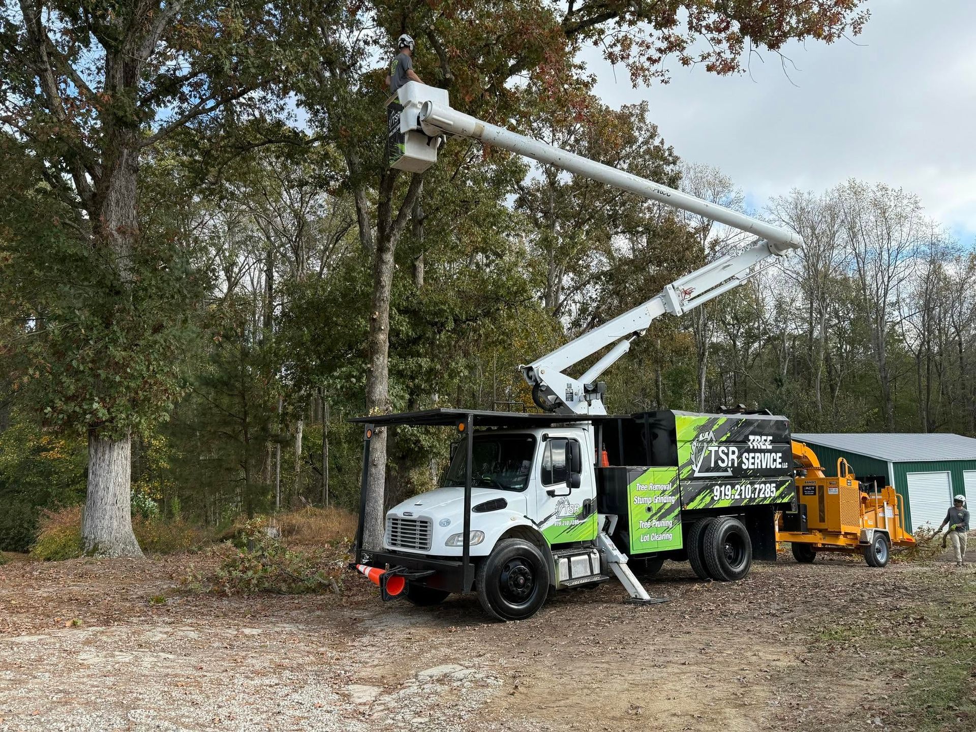 Utility truck with raised bucket and crew in a wooded roadside area, with a yellow wood chipper nearby