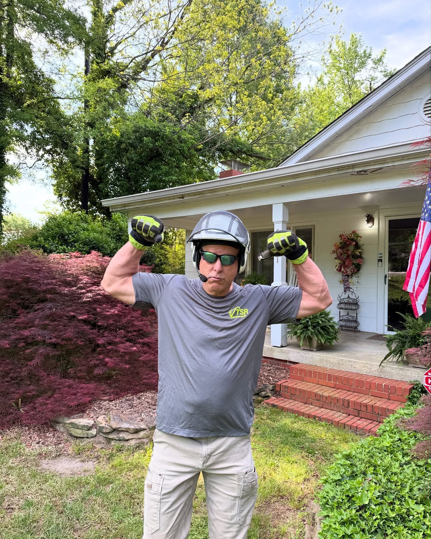Person in helmet and sunglasses flexes in front of a house with green and red landscaping.