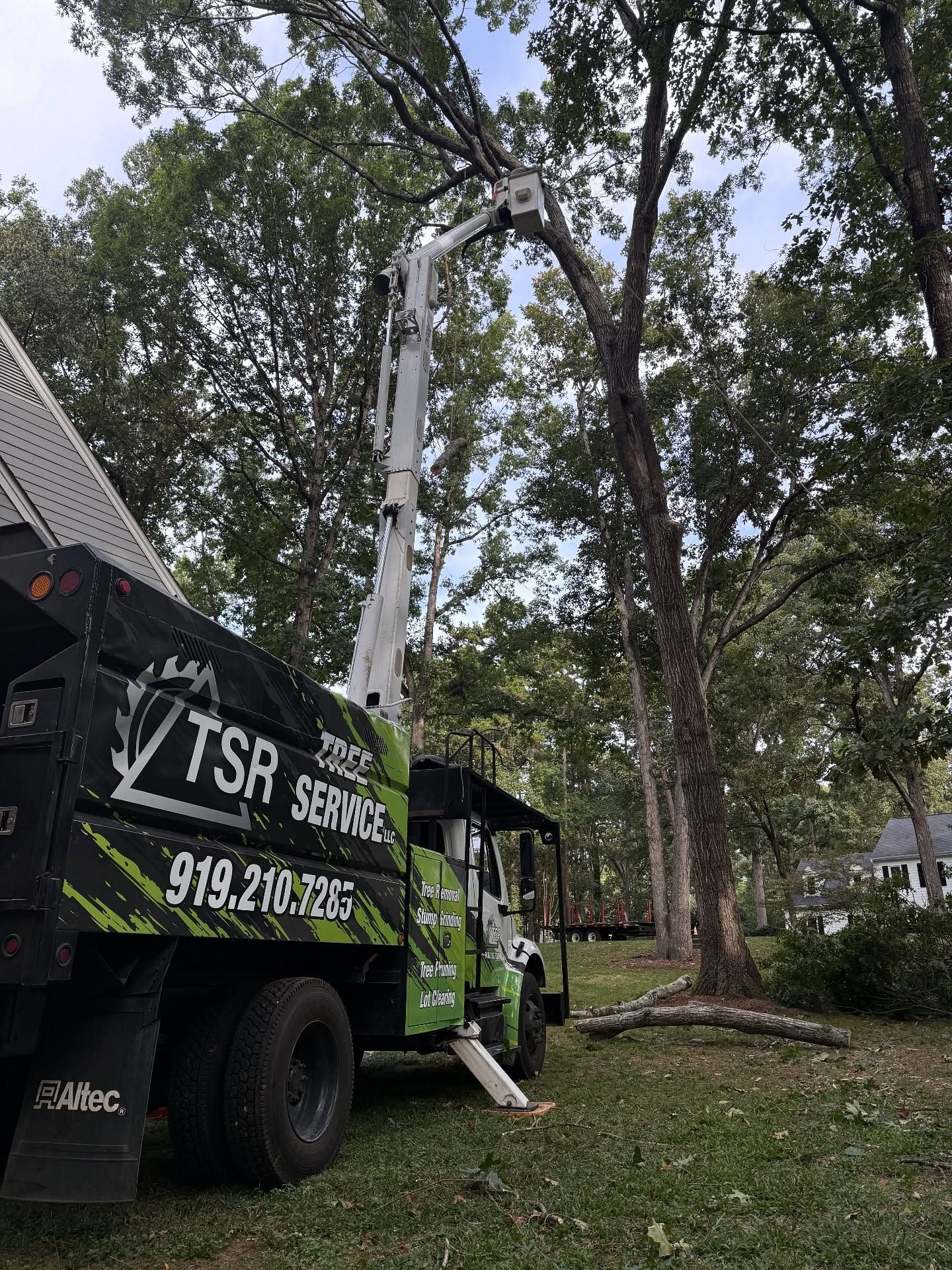 Tree-trimming truck lifting worker beside a large tree in a yard, with fallen branches and a house nearby