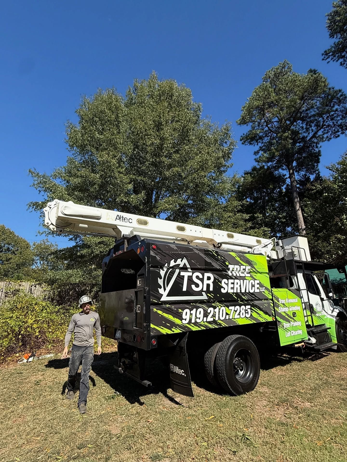 Tree service truck with ladder and worker standing beside it in a sunny grassy yard