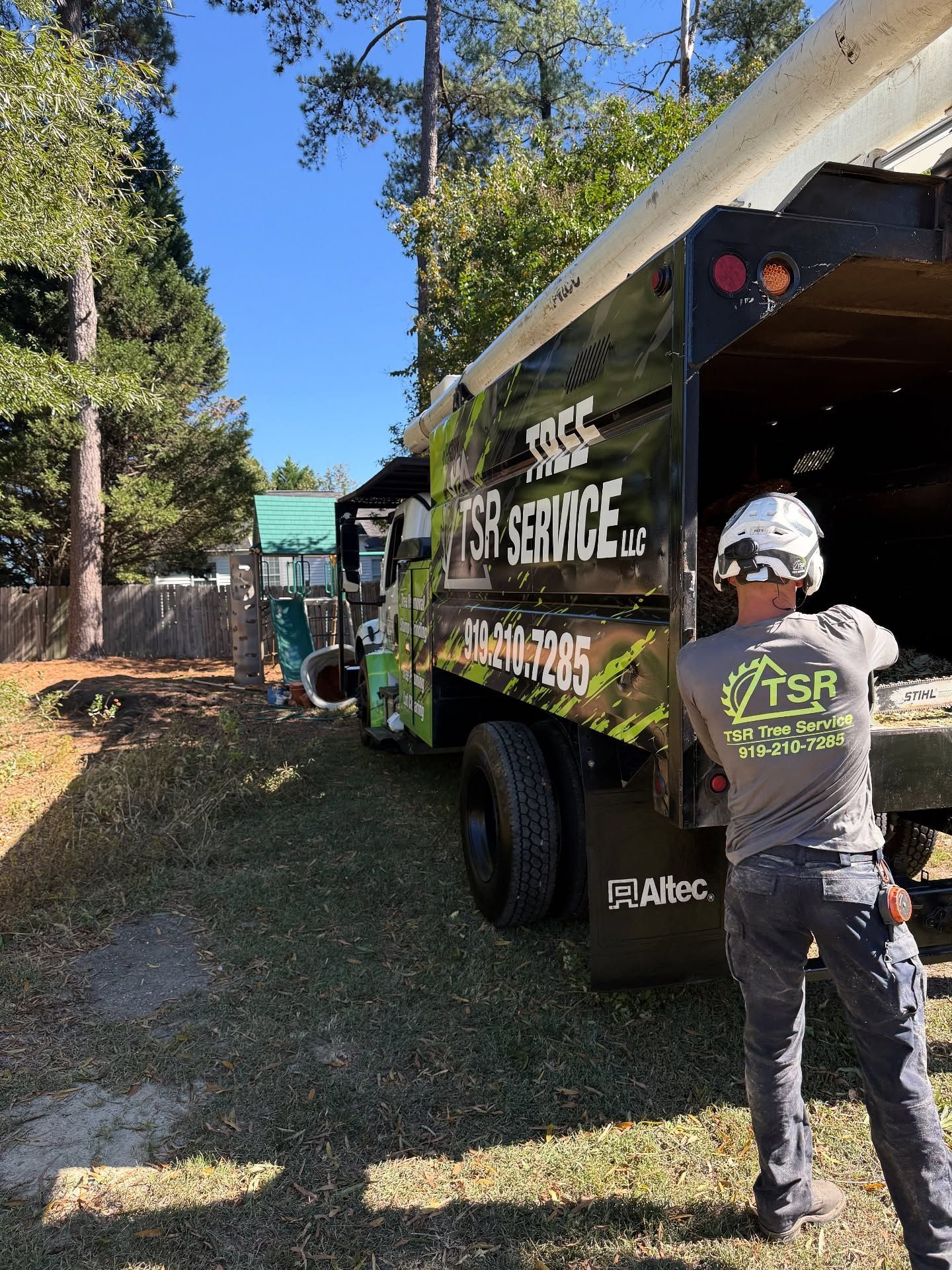 Worker beside green utility truck on a wooded roadside, wearing safety gear and preparing equipment.