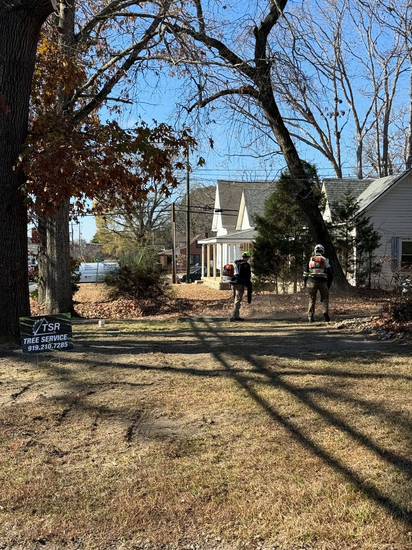 Two people walking on a grassy yard toward a white house under bare trees on a sunny day