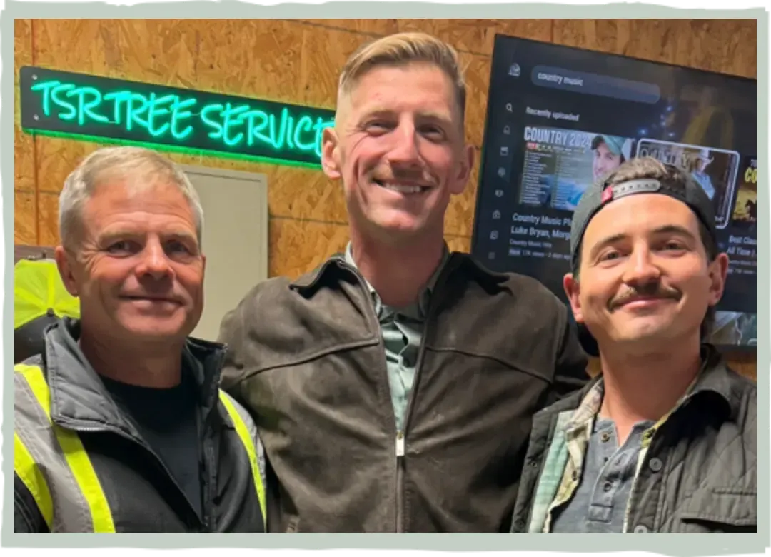 Three men smiling indoors in front of a Tree Service sign and a computer screen.