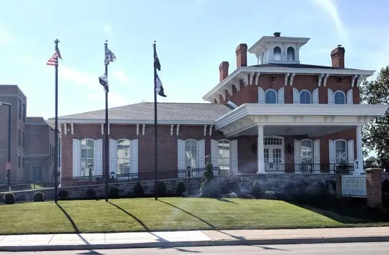 Brick civic building with white trim and columns, two flags, and a manicured lawn on a sunny day