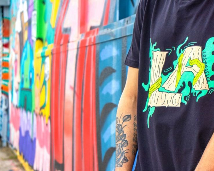 Person in black graphic T-shirt standing by a colorful graffiti wall with tattooed arm visible