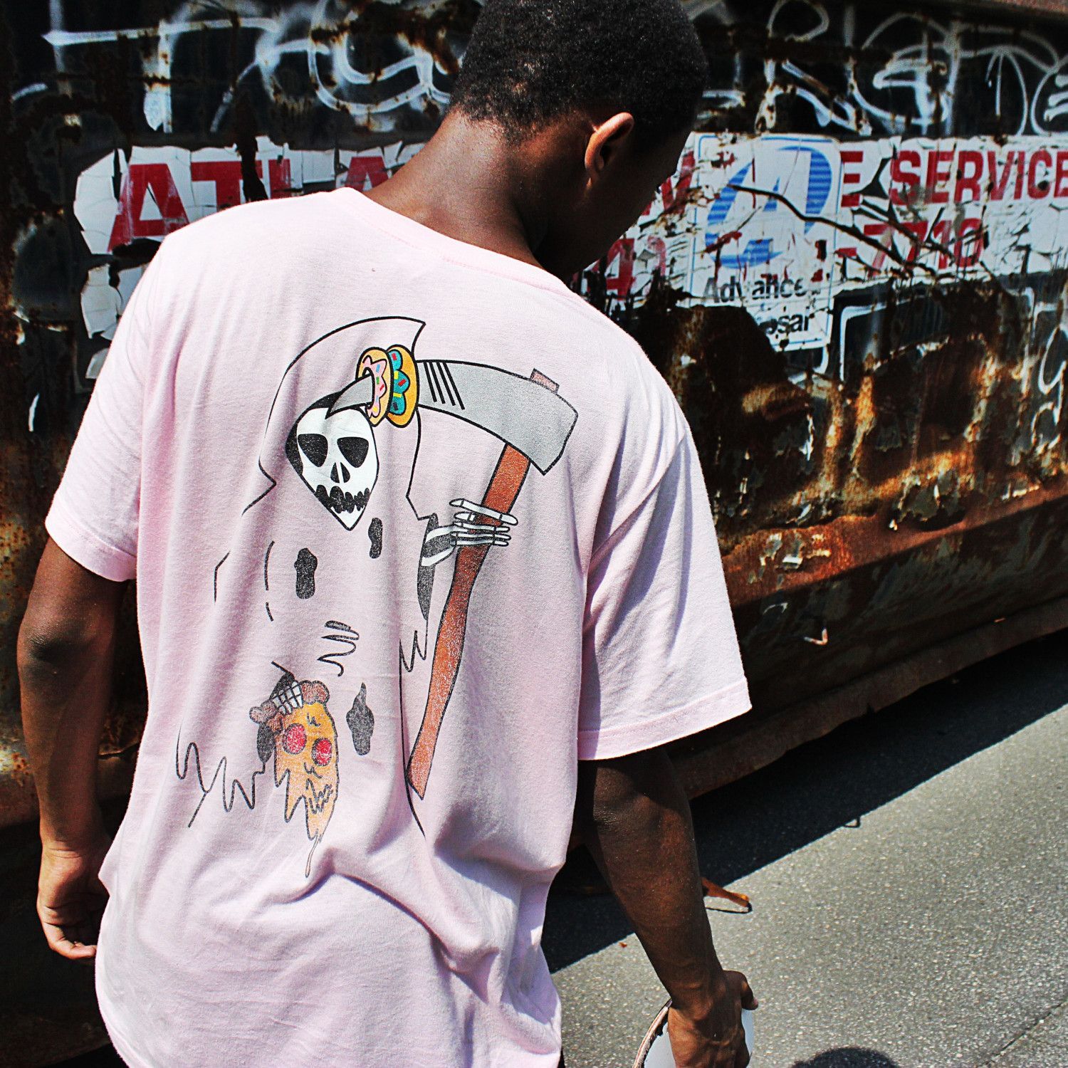 Man in a white T-shirt with a skull graphic standing by a graffiti-covered wall and bus.