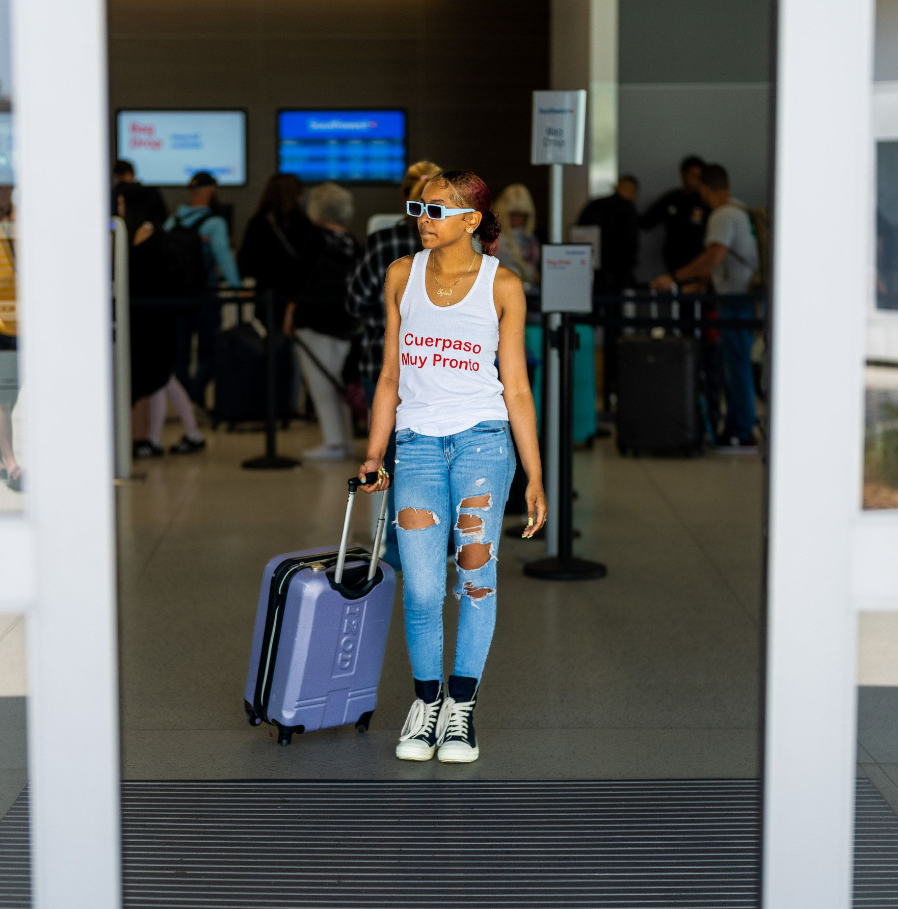 Traveler pulling a purple suitcase through an airport terminal, wearing ripped jeans and a white tank top.