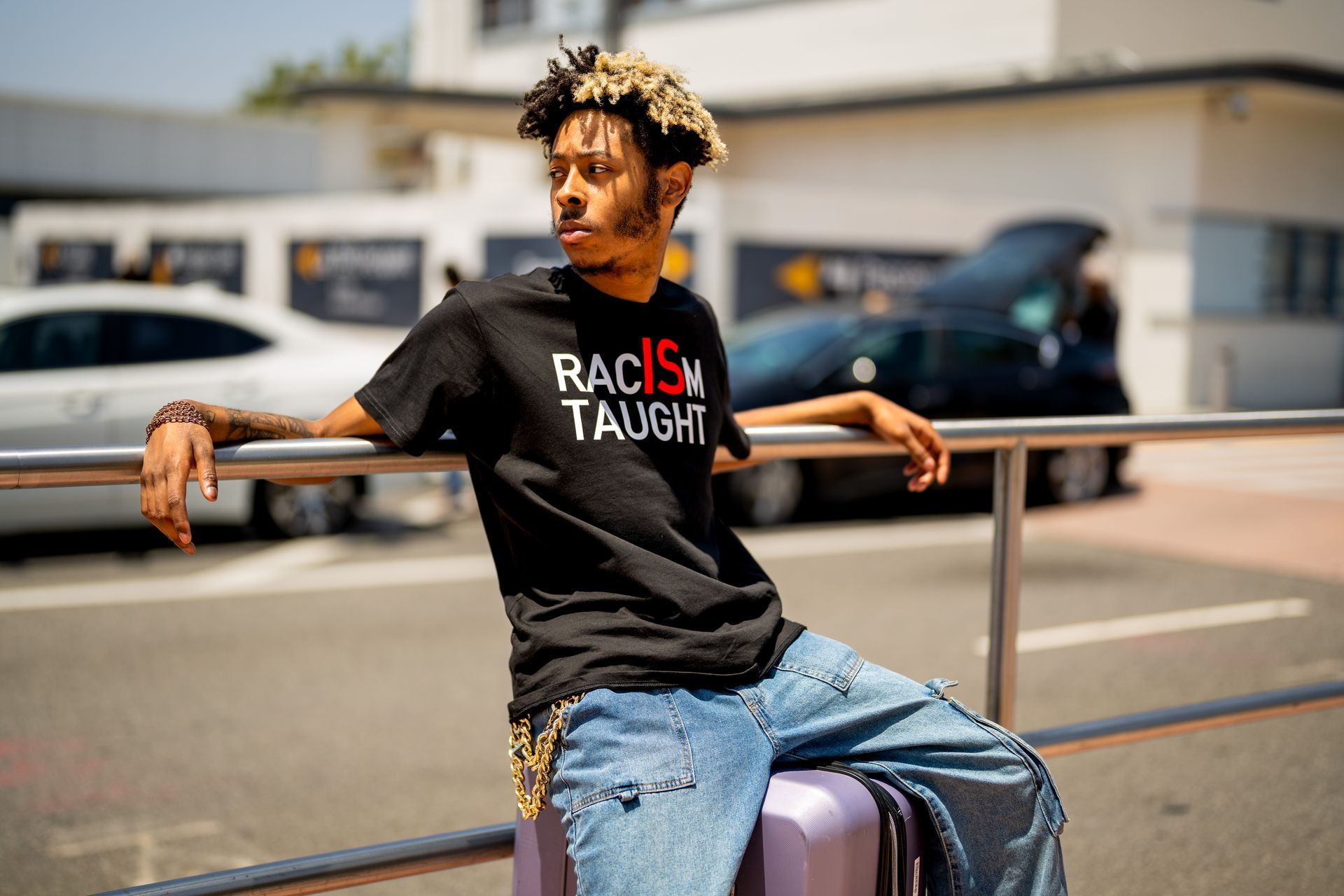 Person in a black shirt and jeans sitting on a suitcase by a rail outdoors, looking left.