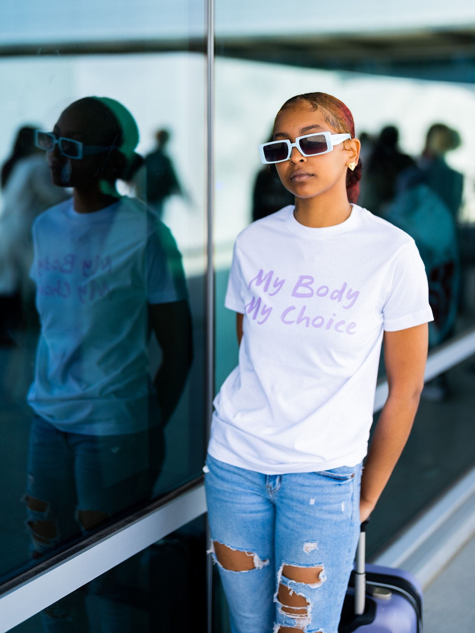 Person in white T-shirt and ripped jeans leaning by a glass wall, with reflections behind them.
