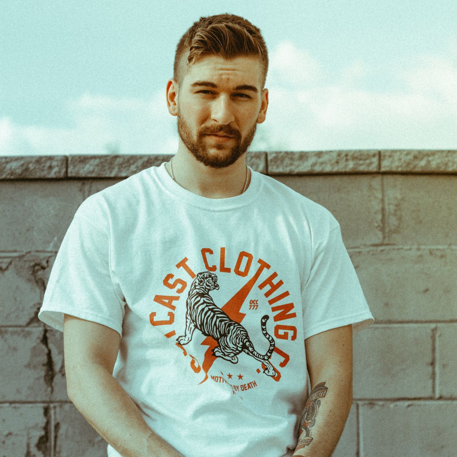 Man standing outdoors against a concrete wall, wearing a white East Clothing T-shirt.