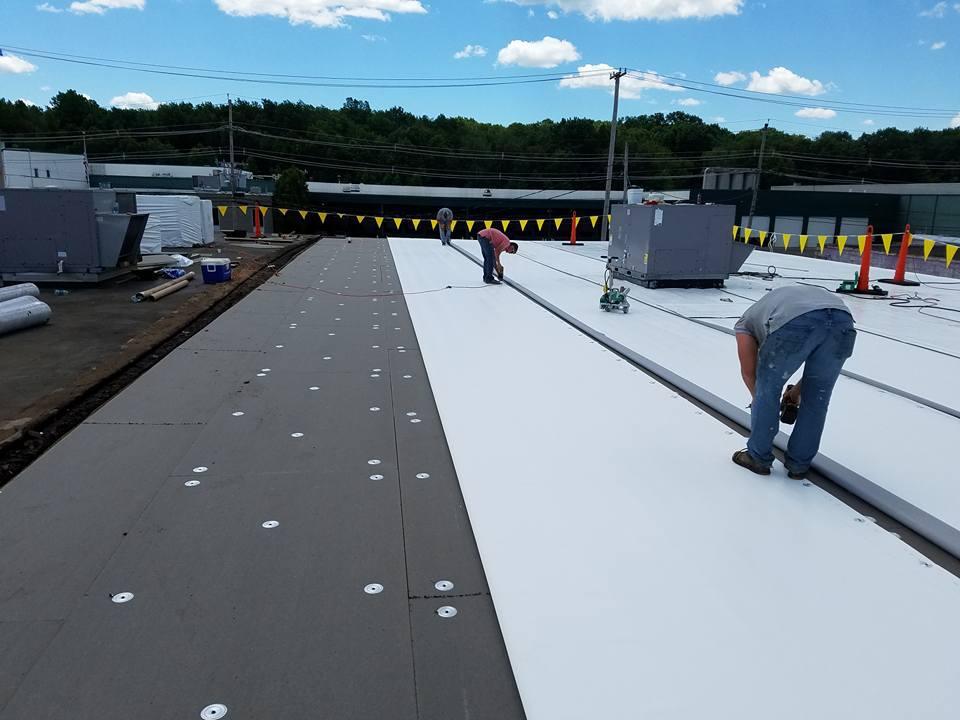 Workers installing white roofing membrane on a flat roof, with tools and construction barriers nearby.