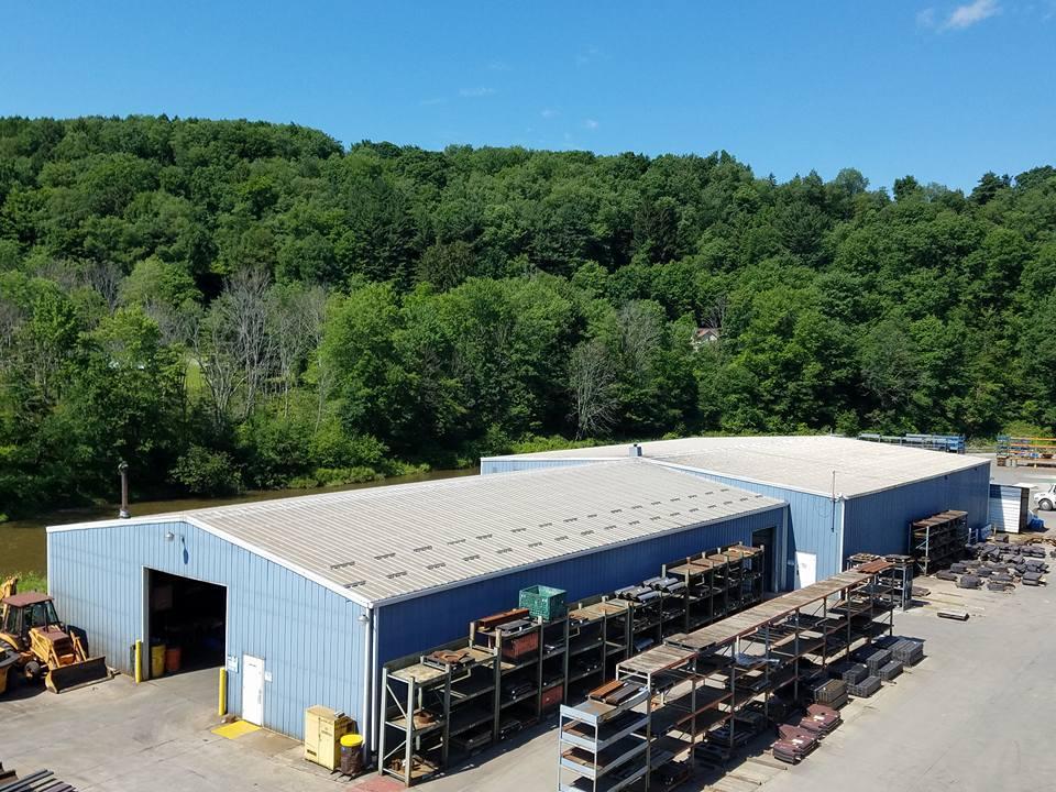 Blue warehouse with outdoor storage racks beside a wooded hillside under a clear sky