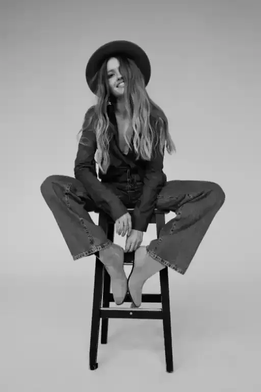 Donna seduta su uno sgabello, con cappello, blazer e jeans, sorridente, a piedi nudi. Ambientazione in studio.