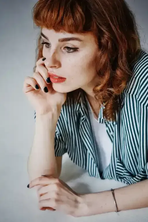Donna con i capelli rossi, il mento appoggiato sulla mano, che indossa una camicia a righe, con aria pensierosa.