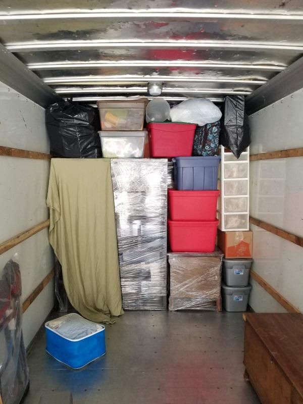 A moving truck interior filled with boxes, bins, and furniture wrapped in plastic, ready for transport.