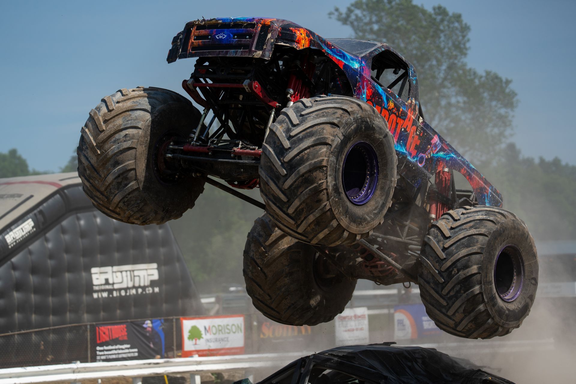 Monster truck with black, red, and blue livery airborne over a dirt track, kicking up dust