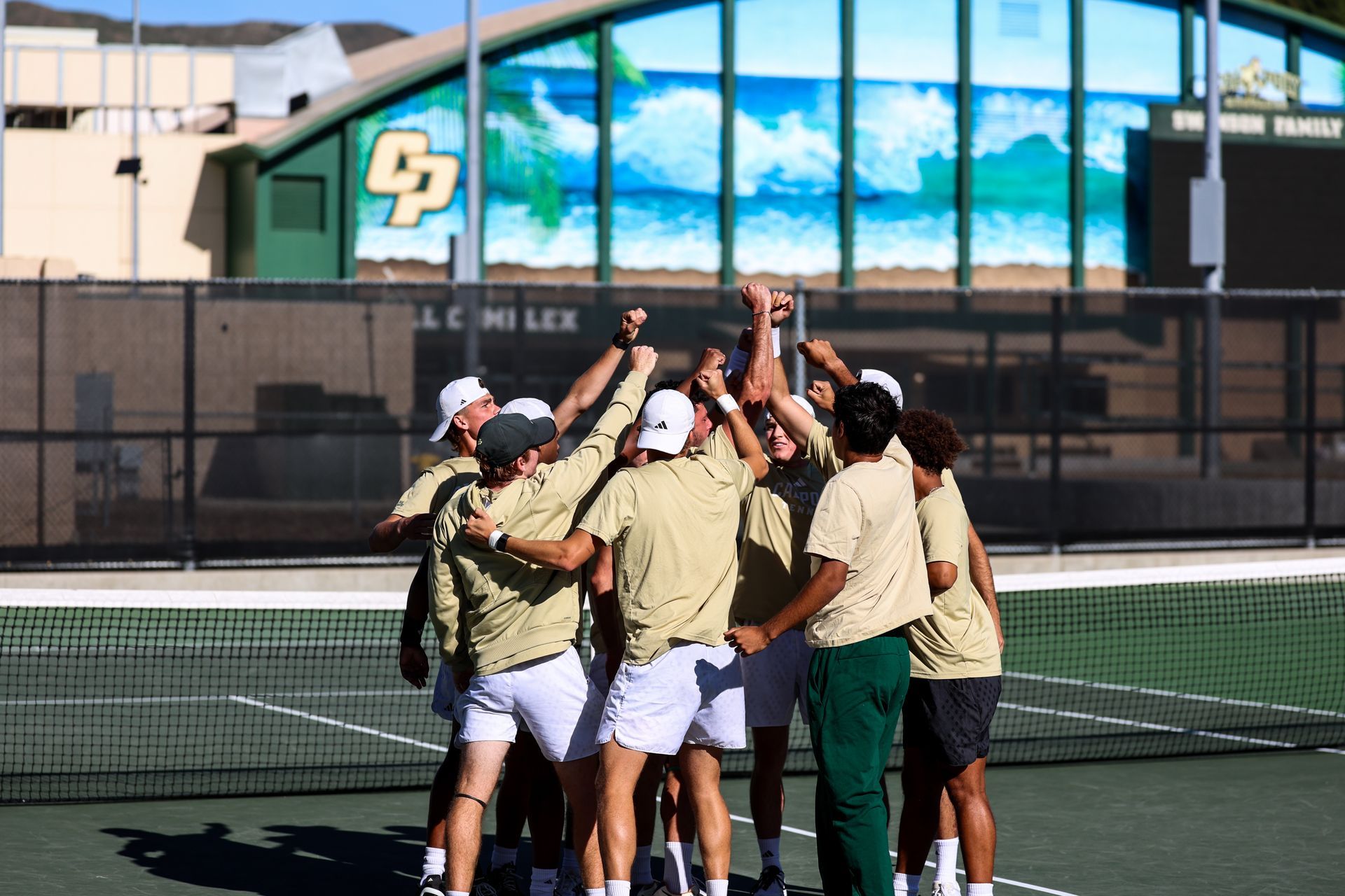 Tennis team huddles with hands raised on an outdoor court in front of a building.