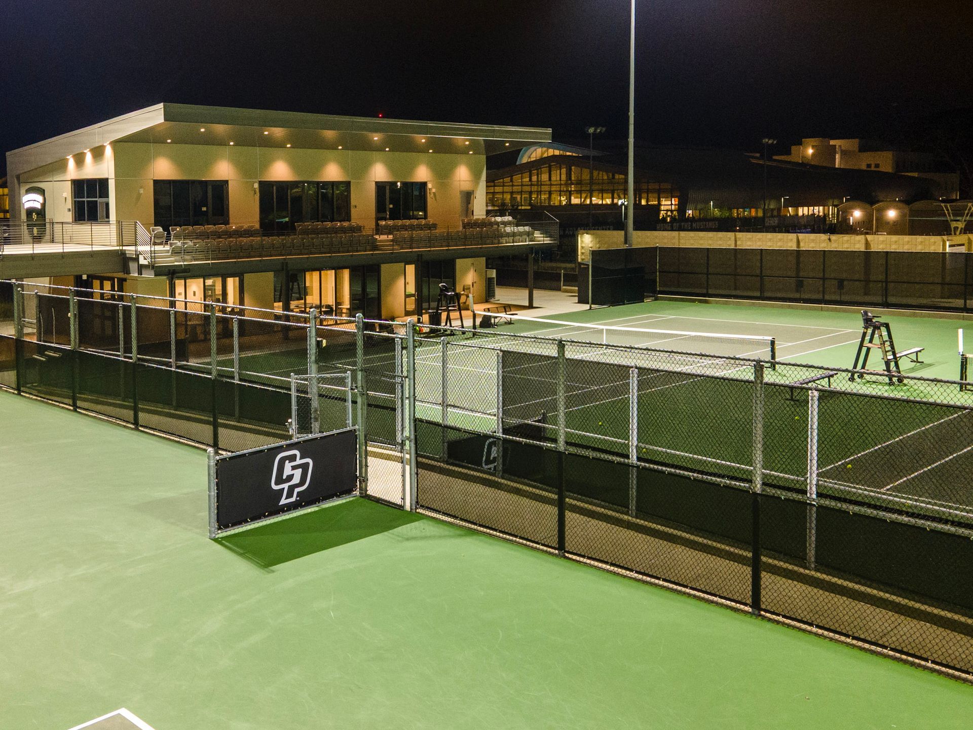 Nighttime outdoor tennis courts with green surfaces and a lit clubhouse behind a chain-link fence