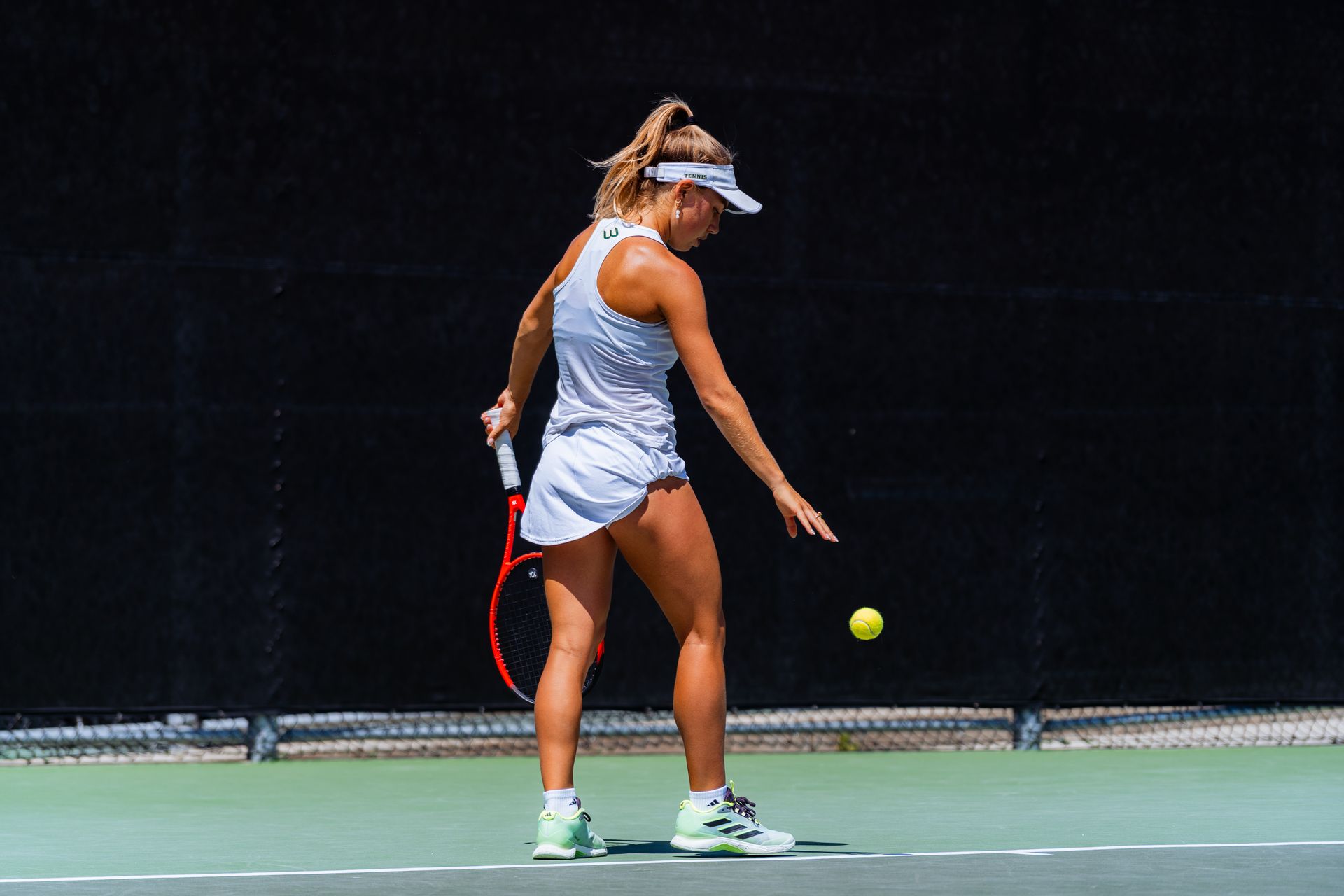 Tennis player in white practice outfit reaches for a low ball on an outdoor court.