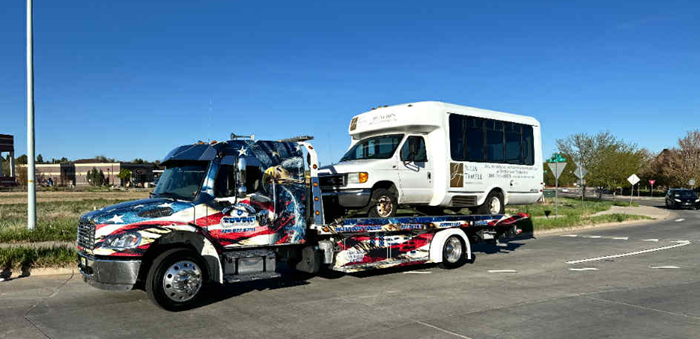 Tow truck carrying a white bus on a sunny day. American flag-themed truck.