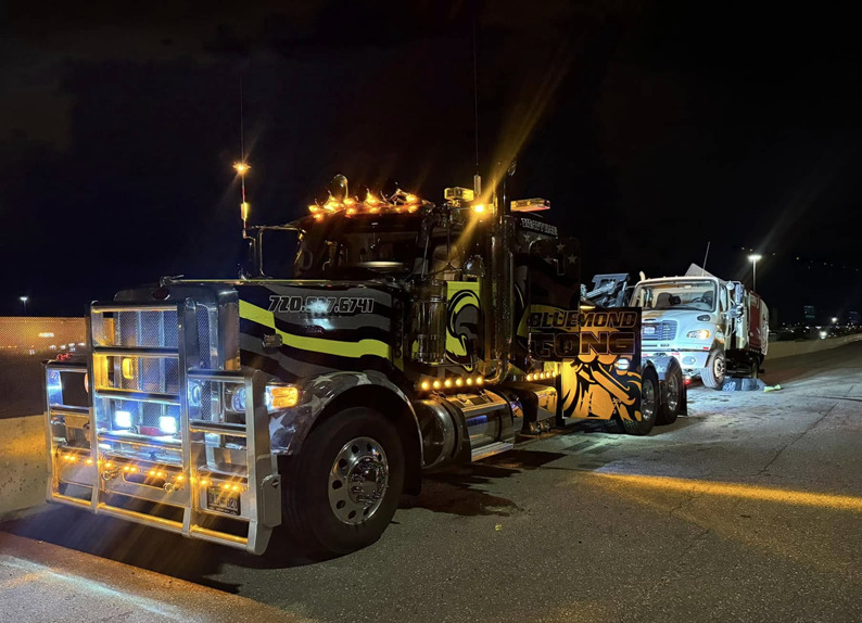 Tow truck with a damaged garbage truck on a dark road at night, lights on.