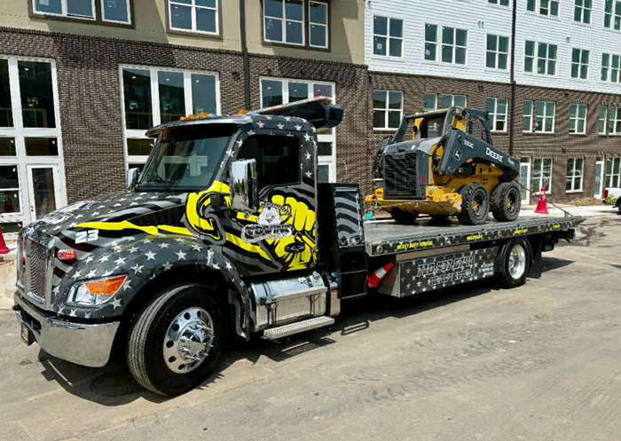 Flatbed tow truck with American flag design, carrying a yellow skid steer at a construction site.