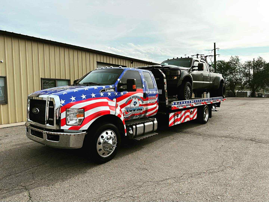 Tow truck with American flag design hauling a dark-colored SUV. Parked outside a building on a sunny day.