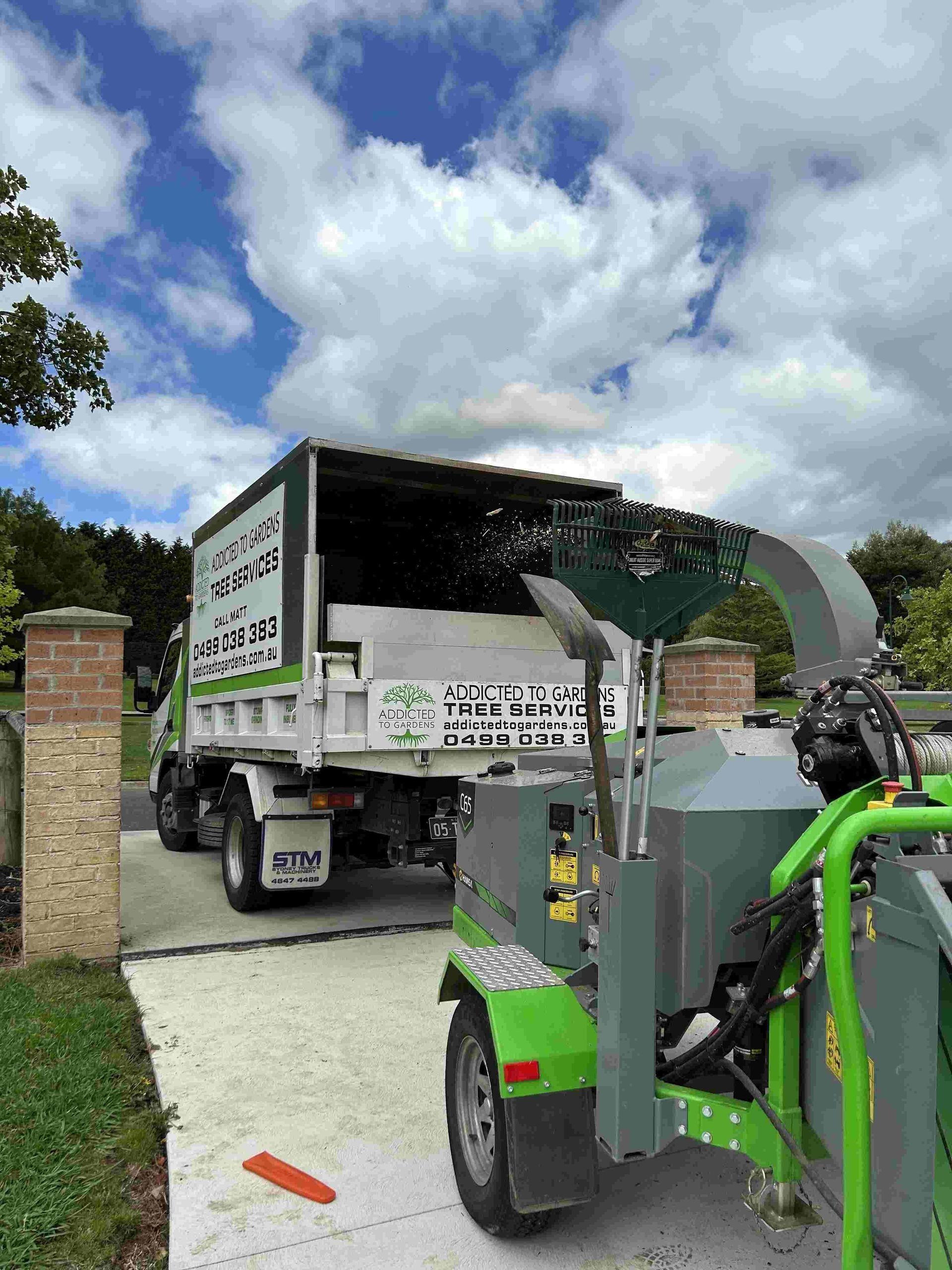 A tree service truck with a wood chipper. The truck is unloading debris under a cloudy sky.
