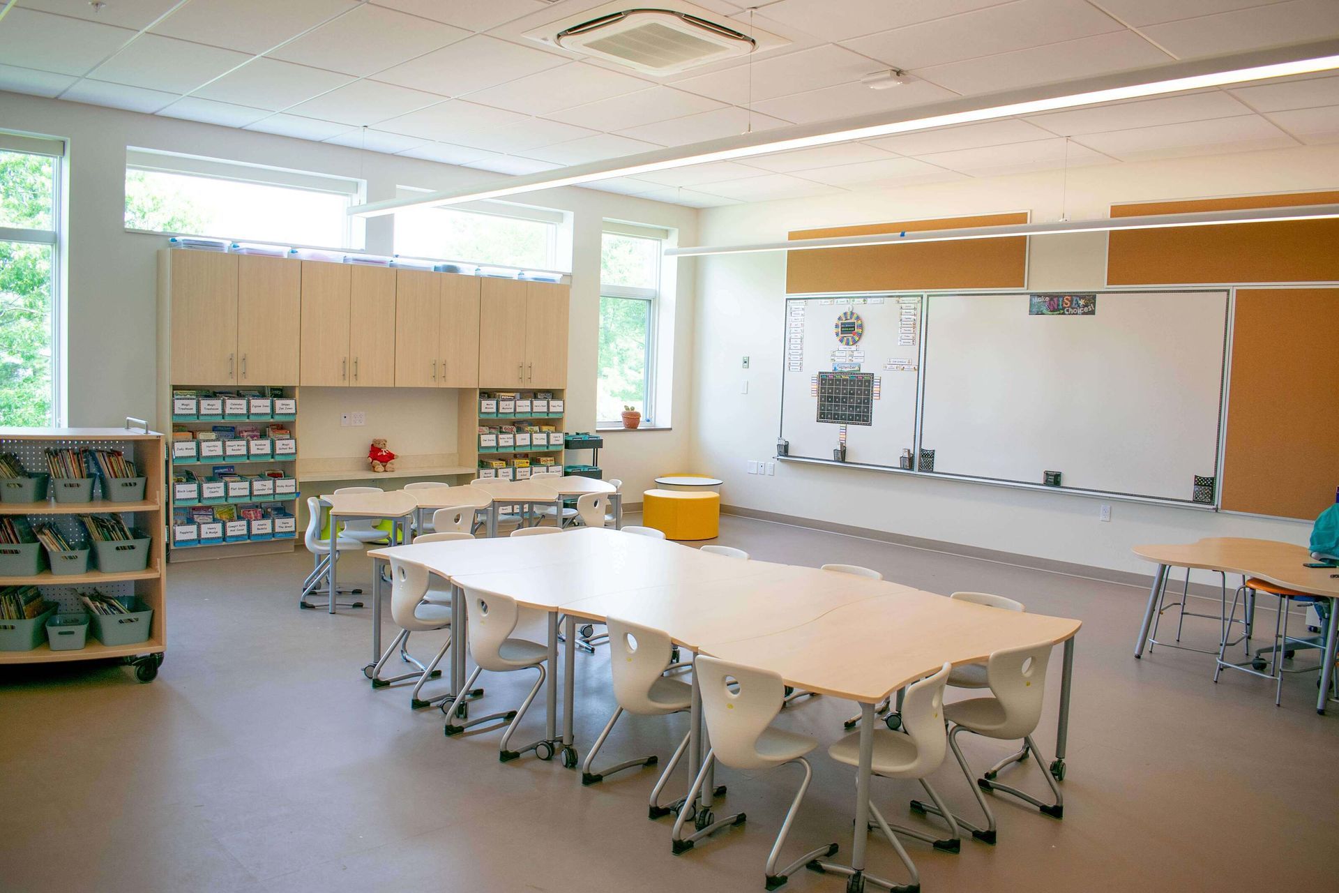 Bright classroom with tables, chairs, cabinets, and a large whiteboard on the wall.