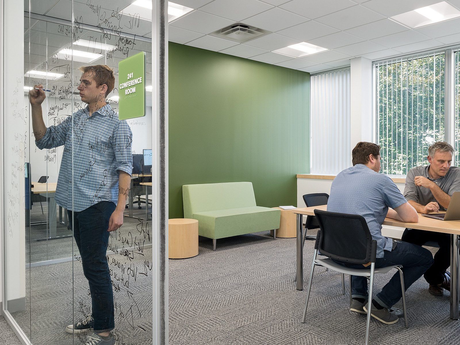 Open office meeting room with a man writing on a glass wall and three people talking at a table