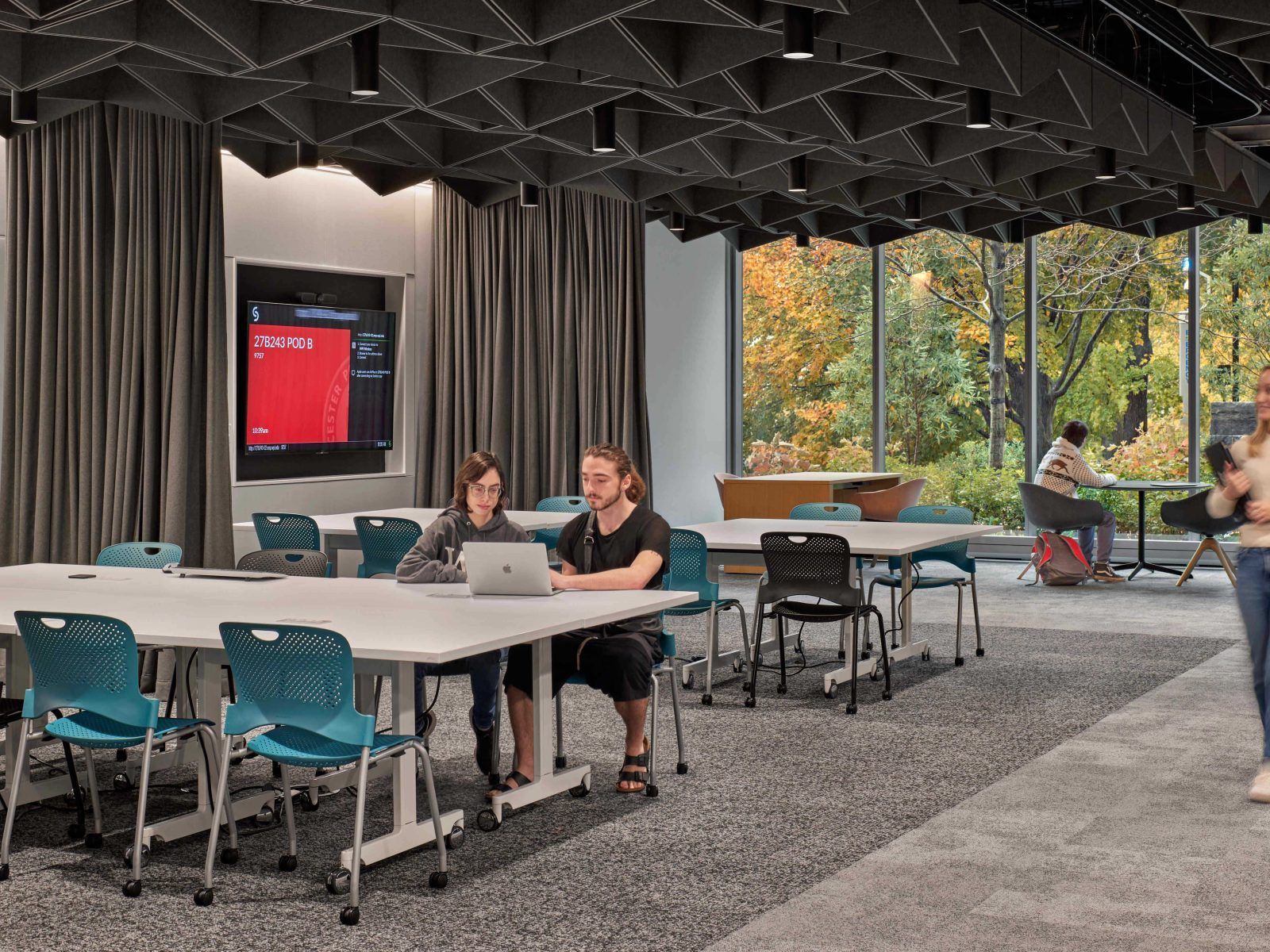 Modern lounge with people at tables, gray curtains, teal chairs, and a red screen by large windows.