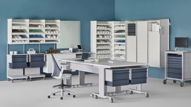 Modern medical office with white desks, blue chairs, shelves, cabinets, and two monitors
