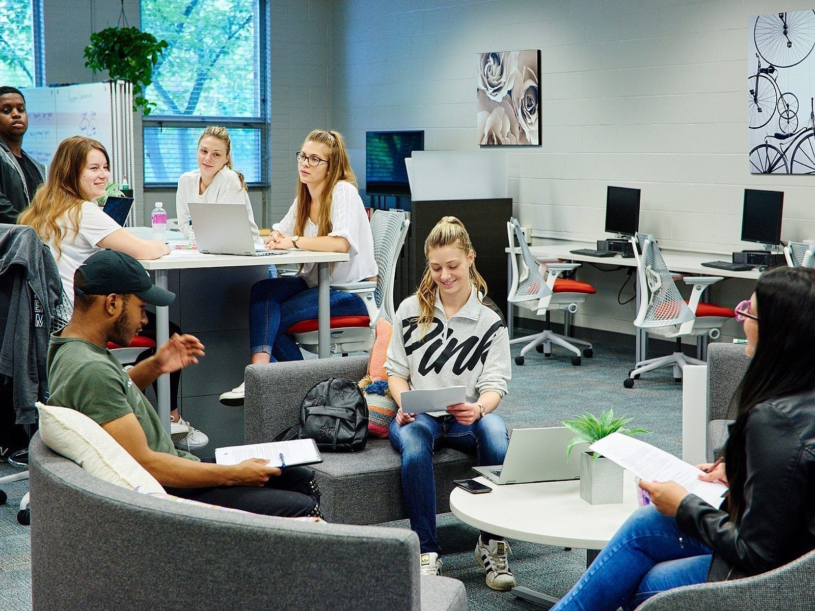 People meeting in a modern office lounge, chatting around tables with laptops and papers.
