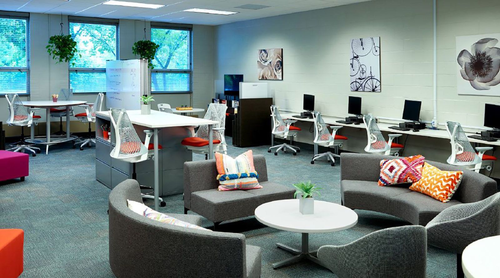 Modern office lounge with gray couches, colorful pillows, desks, and large windows with plants