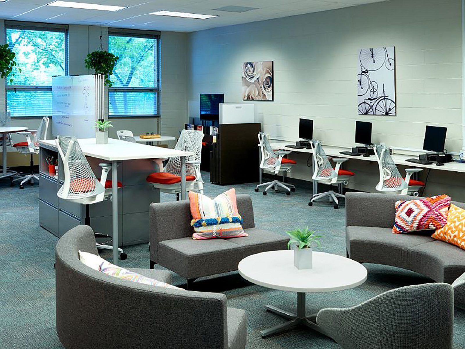 Modern open office lounge with gray seating, round table, desks, computers, and large windows.
