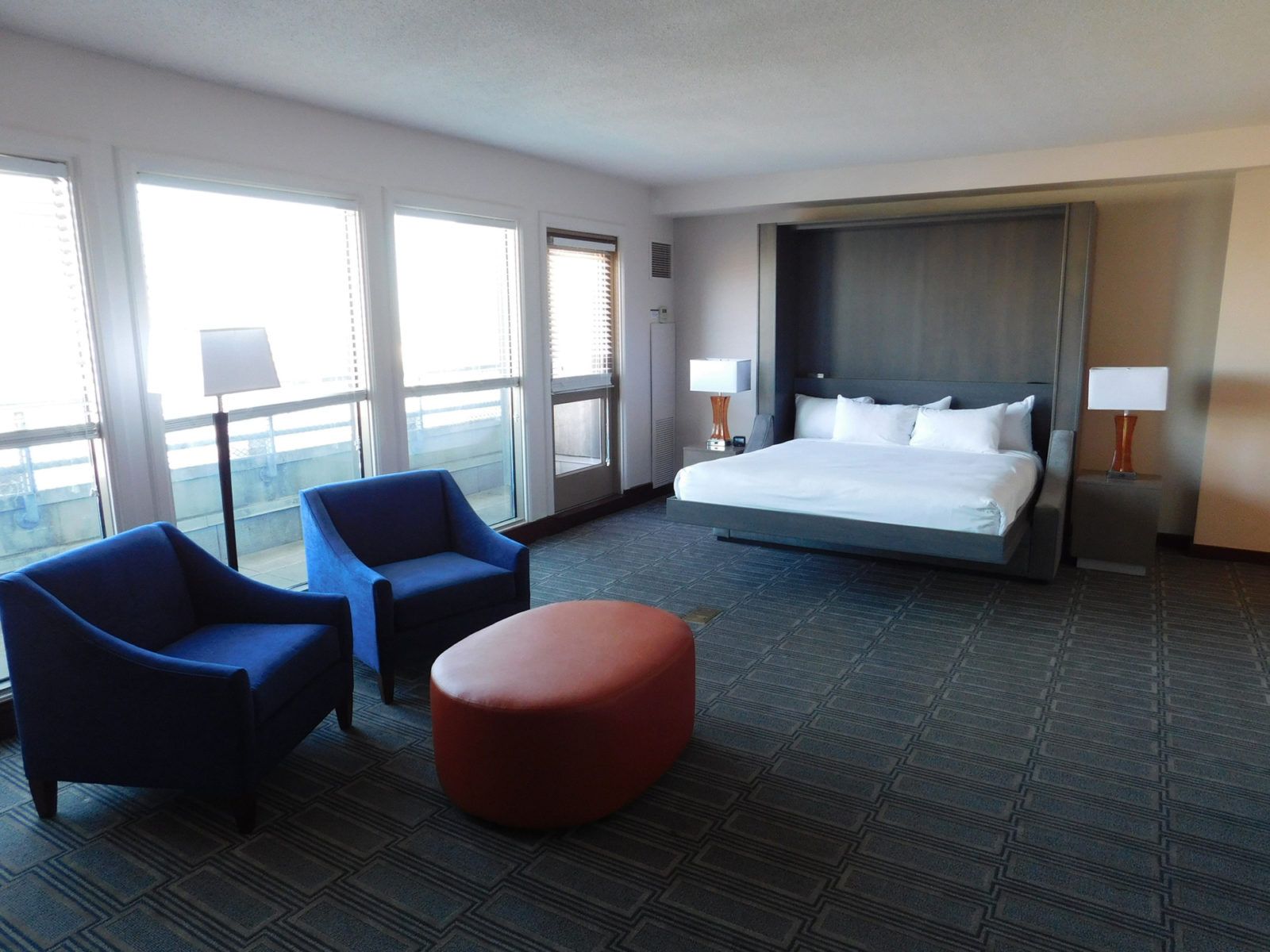 Modern hotel room with a large bed, two blue chairs, and a round orange ottoman by tall windows.