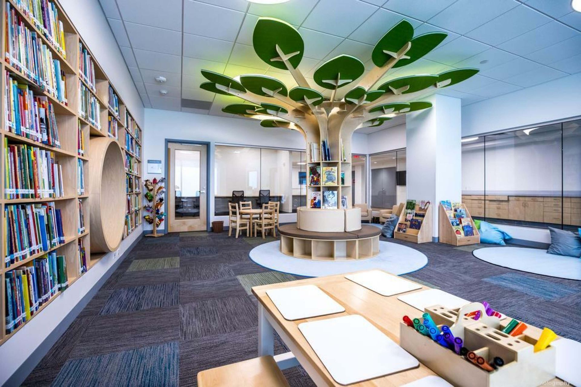 Bright library kids’ area with bookshelves, tree-like seating, and craft table under leafy ceiling decor